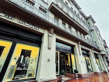 A stylish building with a fashion boutique featuring decorative architectural details. Mannequins displaying suits are visible through large, illuminated windows. The facade has ornamental columns and sculptural elements, with a sign in Cyrillic above the entrance.