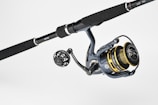 A fishing rod and reel combination featuring a dark handle with metallic accents. The reel has gold and black components with detailed engravings and visible brand markings. It is positioned against a plain, light background.