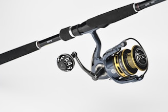 A fishing rod and reel combination featuring a dark handle with metallic accents. The reel has gold and black components with detailed engravings and visible brand markings. It is positioned against a plain, light background.