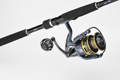 A fishing rod and reel combination featuring a dark handle with metallic accents. The reel has gold and black components with detailed engravings and visible brand markings. It is positioned against a plain, light background.