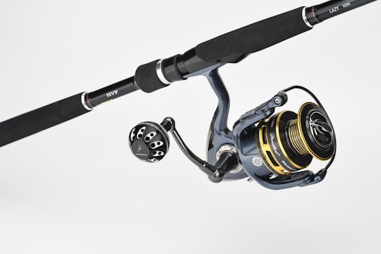 A fishing rod and reel combination featuring a dark handle with metallic accents. The reel has gold and black components with detailed engravings and visible brand markings. It is positioned against a plain, light background.