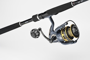 A fishing rod and reel combination featuring a dark handle with metallic accents. The reel has gold and black components with detailed engravings and visible brand markings. It is positioned against a plain, light background.