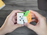 Two hands holding a partially solved Rubik's Cube with green, orange, and white colored squares visible. The cube is branded with a logo. The background is a blurred wooden surface.