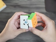 Two hands holding a partially solved Rubik's Cube with green, orange, and white colored squares visible. The cube is branded with a logo. The background is a blurred wooden surface.