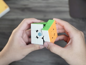 Two hands holding a partially solved Rubik's Cube with green, orange, and white colored squares visible. The cube is branded with a logo. The background is a blurred wooden surface.