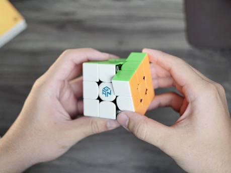 Two hands holding a partially solved Rubik's Cube with green, orange, and white colored squares visible. The cube is branded with a logo. The background is a blurred wooden surface.