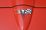 A close-up of a red car hood featuring a chrome emblem in the center. The emblem includes a checkered flag on the right, a v-shaped element in the middle, and a flag with a fleur-de-lis on the left.