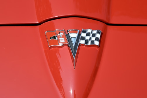 A close-up of a red car hood featuring a chrome emblem in the center. The emblem includes a checkered flag on the right, a v-shaped element in the middle, and a flag with a fleur-de-lis on the left.