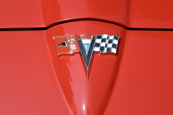 A close-up of a red car hood featuring a chrome emblem in the center. The emblem includes a checkered flag on the right, a v-shaped element in the middle, and a flag with a fleur-de-lis on the left.