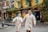 A couple, dressed in matching light beige traditional attire, walks hand in hand down a street lined with yellow-painted buildings and vibrant greenery. Blue shutters adorn the windows of the buildings. Their expressions are joyful, and the street setting suggests a lively, culturally rich environment with colorful lanterns and signs.