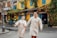 A couple, dressed in matching light beige traditional attire, walks hand in hand down a street lined with yellow-painted buildings and vibrant greenery. Blue shutters adorn the windows of the buildings. Their expressions are joyful, and the street setting suggests a lively, culturally rich environment with colorful lanterns and signs.