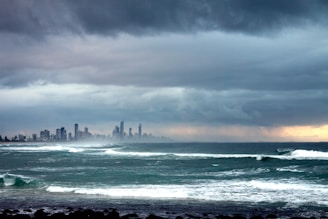 A vast ocean with turbulent waves approaches a distant city skyline under a dramatic, overcast sky. The clouds are dense and dark, with a hint of sunlight peeking through, casting a moody atmosphere over the scene.