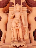 An intricately carved statue of a deity adorned with ornate jewelry and traditional attire, set against a richly detailed backdrop. The figure holds a symbolic object in one hand and has a serene, divine expression.