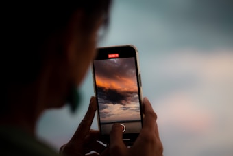 A person holds a smartphone close to their face, capturing a video of a vibrant sky with dramatic clouds. The screen of the phone displays the recording interface with a red 'REC' indicator. The background is blurred, focusing attention on the phone and the scene it is capturing.
