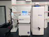 A laboratory setup with a water purification system. The equipment includes a large white reservoir tank and a smaller device with a digital display screen, both labeled with 'Rephile'. There are shelves and power sockets in the background, and various other lab apparatus visible on a counter.