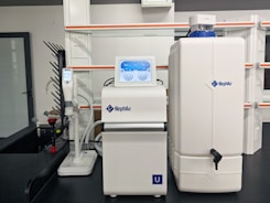 A laboratory setup with a water purification system. The equipment includes a large white reservoir tank and a smaller device with a digital display screen, both labeled with 'Rephile'. There are shelves and power sockets in the background, and various other lab apparatus visible on a counter.