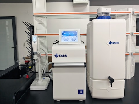 A laboratory setup with a water purification system. The equipment includes a large white reservoir tank and a smaller device with a digital display screen, both labeled with 'Rephile'. There are shelves and power sockets in the background, and various other lab apparatus visible on a counter.