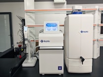 A laboratory setup with a water purification system. The equipment includes a large white reservoir tank and a smaller device with a digital display screen, both labeled with 'Rephile'. There are shelves and power sockets in the background, and various other lab apparatus visible on a counter.