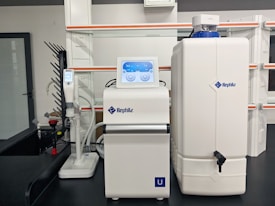 A laboratory setup with a water purification system. The equipment includes a large white reservoir tank and a smaller device with a digital display screen, both labeled with 'Rephile'. There are shelves and power sockets in the background, and various other lab apparatus visible on a counter.