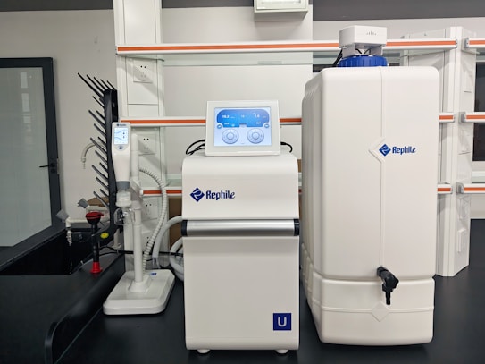 A laboratory setup with a water purification system. The equipment includes a large white reservoir tank and a smaller device with a digital display screen, both labeled with 'Rephile'. There are shelves and power sockets in the background, and various other lab apparatus visible on a counter.