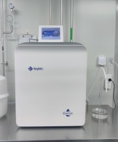 A sleek, modern laboratory water purification system is placed on a stainless steel countertop. The device is predominantly white with a digital control display at the top, featuring the brand logos 'Rephile' and 'Genie'. Surrounding the unit are various laboratory fixtures, including pipes, a pressure gauge, and a clipboard with documentation. A plug with cords is connected on the side, alongside a small transparent container.