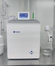 A sleek, modern laboratory water purification system is placed on a stainless steel countertop. The device is predominantly white with a digital control display at the top, featuring the brand logos 'Rephile' and 'Genie'. Surrounding the unit are various laboratory fixtures, including pipes, a pressure gauge, and a clipboard with documentation. A plug with cords is connected on the side, alongside a small transparent container.