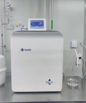A sleek, modern laboratory water purification system is placed on a stainless steel countertop. The device is predominantly white with a digital control display at the top, featuring the brand logos 'Rephile' and 'Genie'. Surrounding the unit are various laboratory fixtures, including pipes, a pressure gauge, and a clipboard with documentation. A plug with cords is connected on the side, alongside a small transparent container.