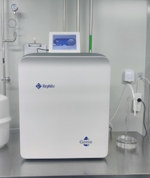 A sleek, modern laboratory water purification system is placed on a stainless steel countertop. The device is predominantly white with a digital control display at the top, featuring the brand logos 'Rephile' and 'Genie'. Surrounding the unit are various laboratory fixtures, including pipes, a pressure gauge, and a clipboard with documentation. A plug with cords is connected on the side, alongside a small transparent container.