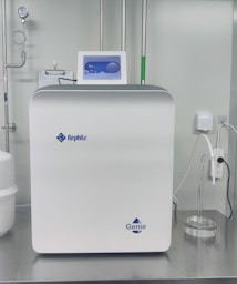 A sleek, modern laboratory water purification system is placed on a stainless steel countertop. The device is predominantly white with a digital control display at the top, featuring the brand logos 'Rephile' and 'Genie'. Surrounding the unit are various laboratory fixtures, including pipes, a pressure gauge, and a clipboard with documentation. A plug with cords is connected on the side, alongside a small transparent container.