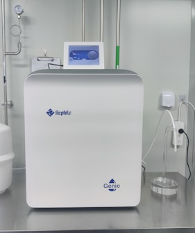 A sleek, modern laboratory water purification system is placed on a stainless steel countertop. The device is predominantly white with a digital control display at the top, featuring the brand logos 'Rephile' and 'Genie'. Surrounding the unit are various laboratory fixtures, including pipes, a pressure gauge, and a clipboard with documentation. A plug with cords is connected on the side, alongside a small transparent container.