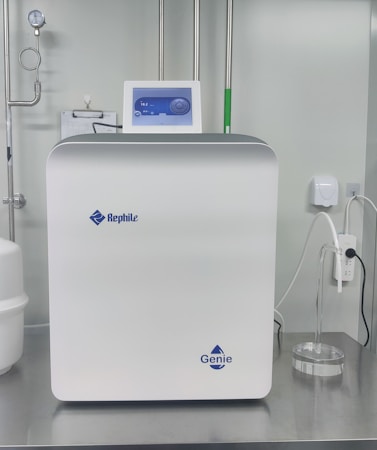 A sleek, modern laboratory water purification system is placed on a stainless steel countertop. The device is predominantly white with a digital control display at the top, featuring the brand logos 'Rephile' and 'Genie'. Surrounding the unit are various laboratory fixtures, including pipes, a pressure gauge, and a clipboard with documentation. A plug with cords is connected on the side, alongside a small transparent container.