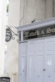 A storefront with closed, gray painted doors is seen, featuring some graffiti and a vintage-style sign above. The sign reads 'Tabak & Sons' and is accompanied by an ornate wrought-iron hanging sign on the side. The environment gives off an urban, slightly aged feel, surrounded by large stone walls.