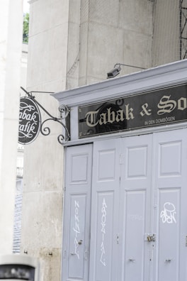 A storefront with closed, gray painted doors is seen, featuring some graffiti and a vintage-style sign above. The sign reads 'Tabak & Sons' and is accompanied by an ornate wrought-iron hanging sign on the side. The environment gives off an urban, slightly aged feel, surrounded by large stone walls.