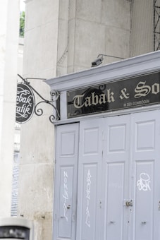 A storefront with closed, gray painted doors is seen, featuring some graffiti and a vintage-style sign above. The sign reads 'Tabak & Sons' and is accompanied by an ornate wrought-iron hanging sign on the side. The environment gives off an urban, slightly aged feel, surrounded by large stone walls.
