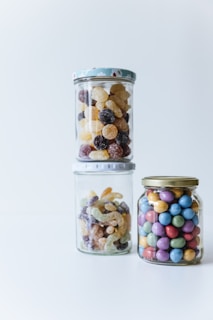 Three glass jars filled with colorful candies are arranged on a white surface. The left and center jars contain various gummy candies in different shapes and colors, while the right jar is filled with round, multicolored coated candies.