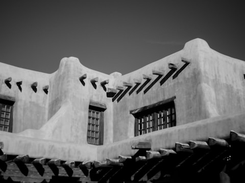A black and white photograph of an adobe-style building featuring stucco walls and wooden beams protruding from the roofline. The structure has a series of small, rectangular windows and exhibits a smooth, rounded architectural design characteristic of Southwestern architecture.