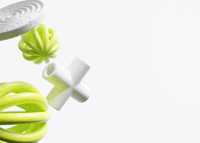 A collection of abstract 3D geometric shapes, including a white spiral disc, a green twisted torus, a spherical green object with a petal-like structure, and a white cross-like form. These shapes have a smooth, glossy texture with small black speckles and are set against a plain light background.
