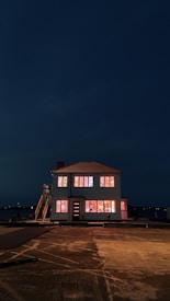 A house with bright, colorful lights illuminating the windows stands against a dark, night sky. The lights create a warm and inviting glow despite the surrounding darkness. The house is positioned near a body of water, adding to the serene atmosphere.