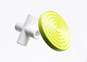 Two abstract geometric objects appear suspended against a light background. One object, resembling a white cylindrical cross, is speckled with small dots. The other is a vibrant yellow disc with concentric ridges, also dotted with small specks.