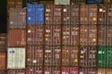 A large stack of shipping containers with various colors, including red, blue, green, and white. Each container is marked with white letters and symbols. The containers are arranged in rows, creating a geometric pattern.