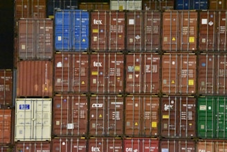 A large stack of shipping containers with various colors, including red, blue, green, and white. Each container is marked with white letters and symbols. The containers are arranged in rows, creating a geometric pattern.