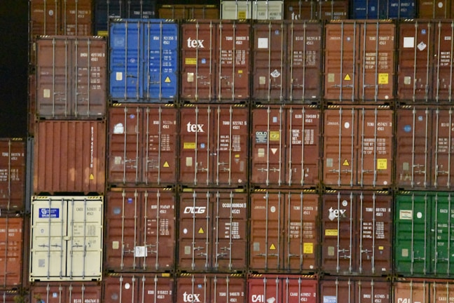 A large stack of shipping containers with various colors, including red, blue, green, and white. Each container is marked with white letters and symbols. The containers are arranged in rows, creating a geometric pattern.