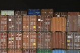 Multiple shipping containers stacked in rows. The containers are of different colors including brown, red, orange, blue, and green, each labeled with various text and symbols. The background is dark, emphasizing the containers.