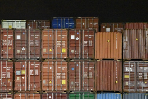 Multiple shipping containers stacked in rows. The containers are of different colors including brown, red, orange, blue, and green, each labeled with various text and symbols. The background is dark, emphasizing the containers.