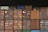 Multiple shipping containers stacked in rows. The containers are of different colors including brown, red, orange, blue, and green, each labeled with various text and symbols. The background is dark, emphasizing the containers.
