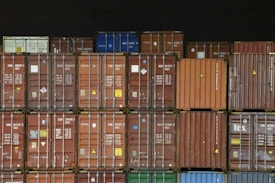 Multiple shipping containers stacked in rows. The containers are of different colors including brown, red, orange, blue, and green, each labeled with various text and symbols. The background is dark, emphasizing the containers.