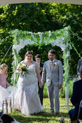 A couple is walking hand in hand down the aisle under an arch covered with greenery and white flowers. The bride is wearing a white lace dress and holding a bouquet of flowers. The groom is dressed in a light grey suit with a white boutonniere. There is a person officiating the ceremony and a young girl in a pink dress following them. The background is lush with green trees and foliage.