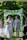 A couple is walking hand in hand down the aisle under an arch covered with greenery and white flowers. The bride is wearing a white lace dress and holding a bouquet of flowers. The groom is dressed in a light grey suit with a white boutonniere. There is a person officiating the ceremony and a young girl in a pink dress following them. The background is lush with green trees and foliage.