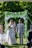 A couple is walking hand in hand down the aisle under an arch covered with greenery and white flowers. The bride is wearing a white lace dress and holding a bouquet of flowers. The groom is dressed in a light grey suit with a white boutonniere. There is a person officiating the ceremony and a young girl in a pink dress following them. The background is lush with green trees and foliage.