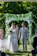 A couple is walking hand in hand down the aisle under an arch covered with greenery and white flowers. The bride is wearing a white lace dress and holding a bouquet of flowers. The groom is dressed in a light grey suit with a white boutonniere. There is a person officiating the ceremony and a young girl in a pink dress following them. The background is lush with green trees and foliage.